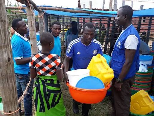 Assorted refugee supply kits – dignity kits, hygiene packs, shelter tarpaulins, jerry cans, and mosquito nets arranged on a pallet, ready for distribution in Uganda.